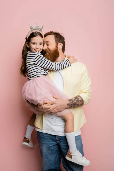 Tattooed man holding cheerful daughter with crown headband on pink background — Stock Photo