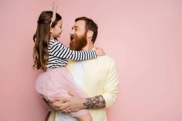 Positive tattooed man holding daughter with crown headband on pink background — Stock Photo