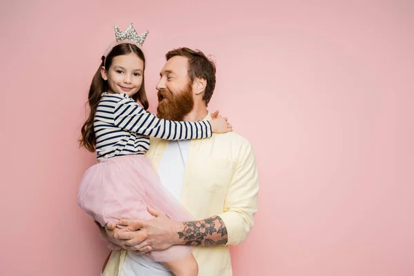 Alegre padre barbudo sosteniendo hija con diadema de corona sobre fondo rosa - foto de stock