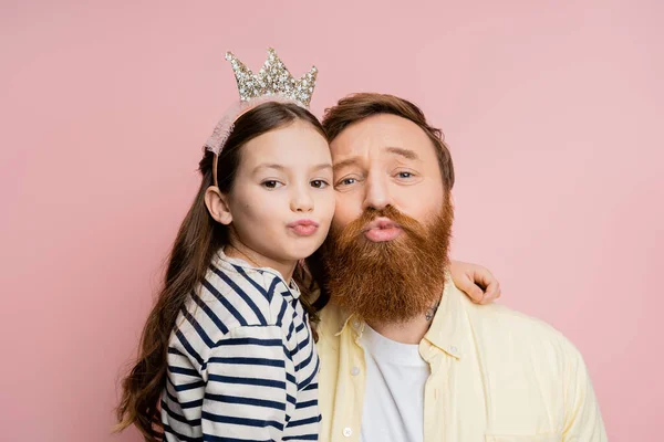 Hija preadolescente con diadema de corona haciendo pucheros labios y abrazando a papá aislado en rosa - foto de stock