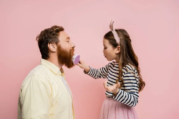 Side view of preteen girl with crown headband brushing beard of father isolated on pink — Stock Photo