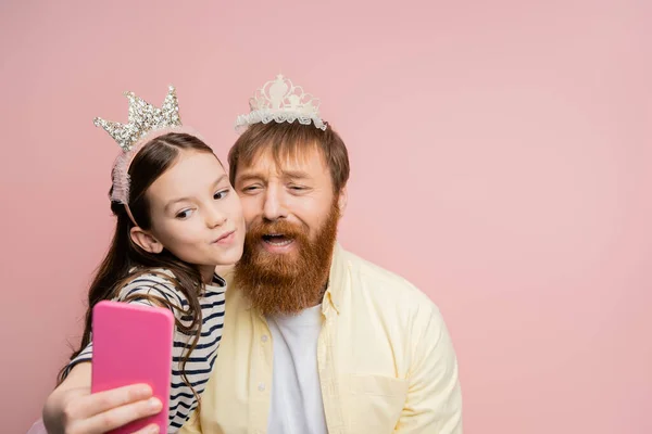 Preteen girl taking selfie with sad dad with crown headband isolated on pink — Stock Photo