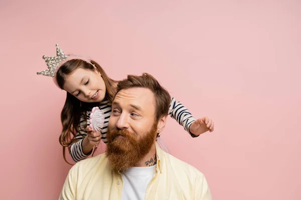 Усміхнена дівчина з коронкою на голові тримає щітку біля бородатого батька ізольовано на рожевому — стокове фото