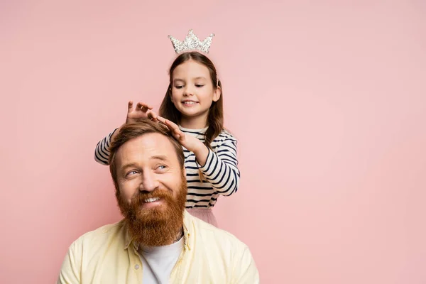 Fröhliches Mädchen mit Krone Stirnband berührt Haar des lächelnden Papas isoliert auf rosa — Stockfoto