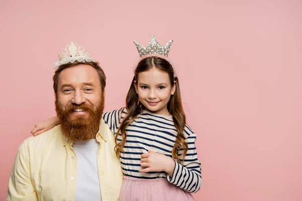 Усміхнена дочка і батько в коронних головних уборах дивиться на камеру ізольовано на рожевому — стокове фото