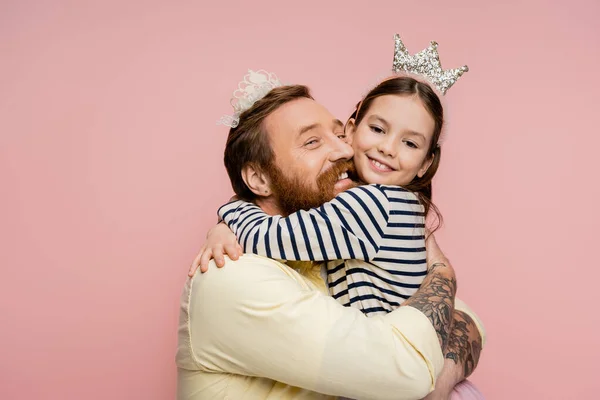 Усміхнений татуйований батько обіймає дочку в короні ізольовано на рожевому — стокове фото