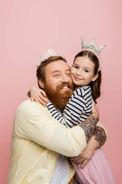 Alegre padre e hija en coronas abrazo aislado en rosa - foto de stock