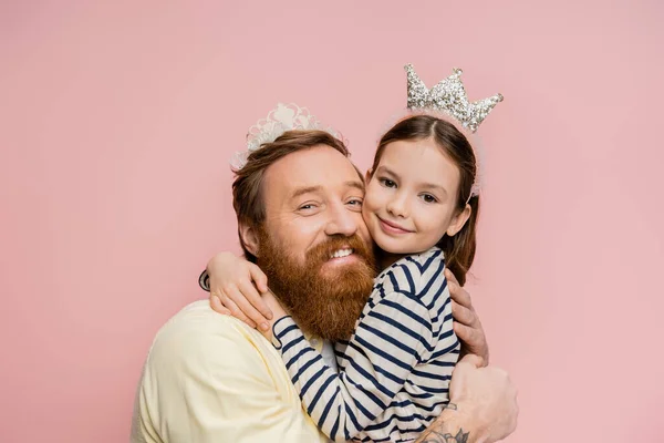 Positive father and preteen girl in crown headbands hugging isolated on pink — Stock Photo
