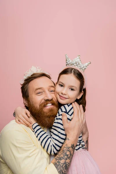 Smiling tattooed man in crown headband hugging daughter isolated on pink — Stock Photo