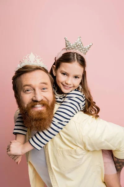 Chica positiva piggybacking en el padre con diadema de corona aislado en rosa - foto de stock
