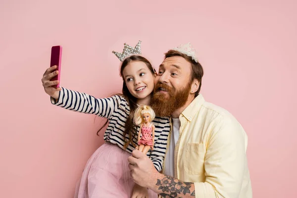 Cheerful preteen girl taking selfie with father with crown headband holding doll on pink background — Stock Photo