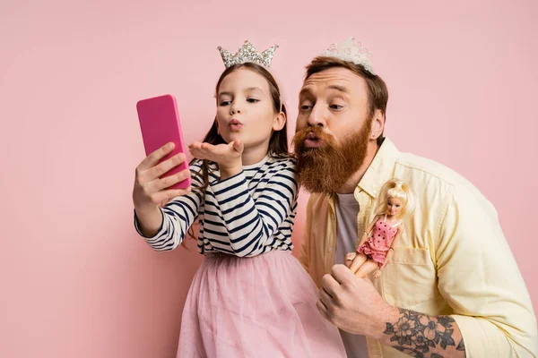 Hija y padre con muñecas y diademas de corona tomando selfie en el teléfono inteligente sobre fondo rosa - foto de stock