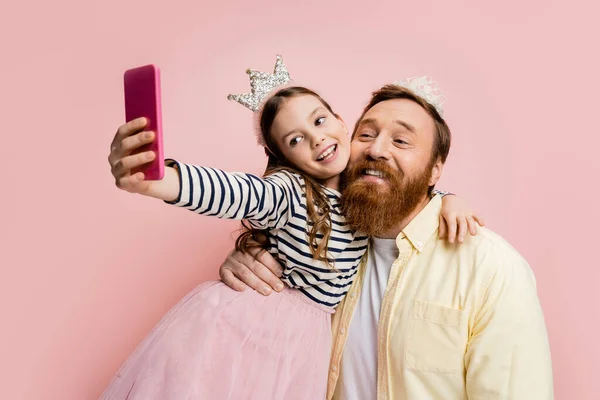 Sonriente chica abrazando padre en diadema de corona y tomando selfie aislado en rosa - foto de stock