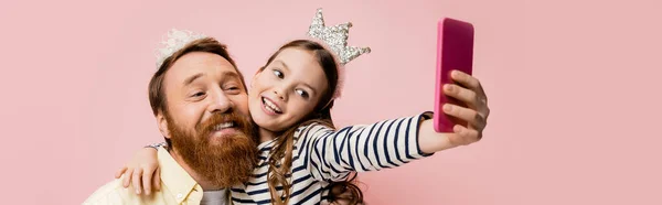 Positive child hugging father with crown headband while taking selfie isolated on pink, banner — Stock Photo