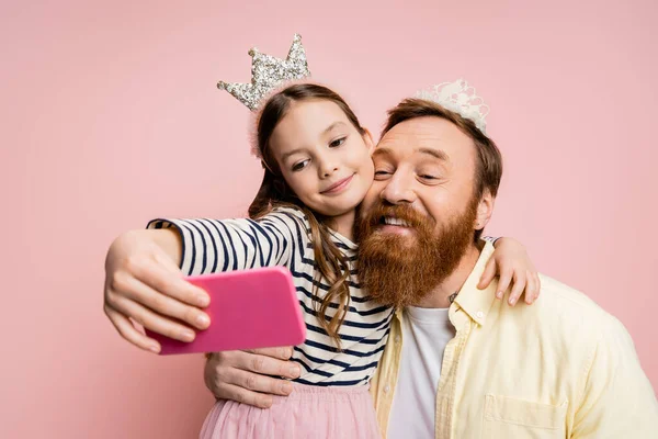 Homme positif avec bandeau couronne prenant selfie avec fille isolée sur rose — Photo de stock