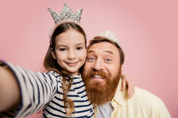 Lächelndes Kind umarmt Vater mit Kronenband und blickt isoliert in die Kamera auf rosa — Stockfoto