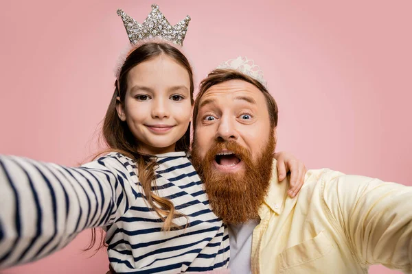 Positive Mann und Tochter mit Krone Stirnbänder Blick in die Kamera isoliert auf rosa — Stockfoto