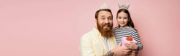 Hombre e hija positivos con diademas de corona sosteniendo regalo aislado en rosa, bandera - foto de stock