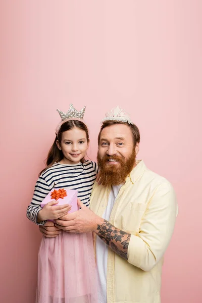 Позитивный мужчина с повязкой на голове с подарочной коробкой рядом с дочерью-подростком на розовом фоне — стоковое фото