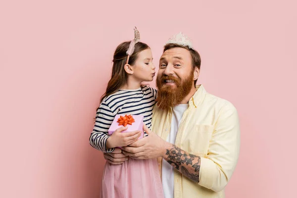 Preteen girl with crown headband holding gift and kissing bearded father on pink background — Stock Photo