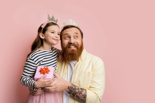 Sonriente hija y papá con diademas de corona sosteniendo caja de regalo sobre fondo rosa - foto de stock