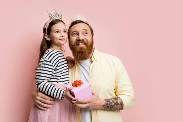 Positive Vater und Tochter mit Krone Stirnbänder halten Geschenk und schauen weg auf rosa Hintergrund — Stockfoto