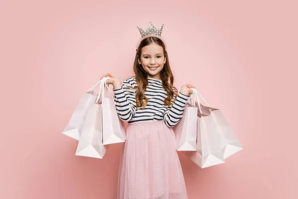 Muchacha preadolescente sonriente en diadema de corona sosteniendo bolsas de compras sobre fondo rosa - foto de stock