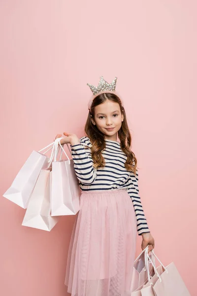 Carefree preteen kid in crown headband holding shopping bags on pink background — Stock Photo