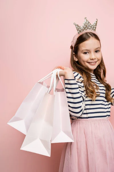 Fröhliches Frühchen im Kronenstirnband hält Einkaufstüten in der Hand und blickt in die Kamera auf rosa Hintergrund — Stockfoto