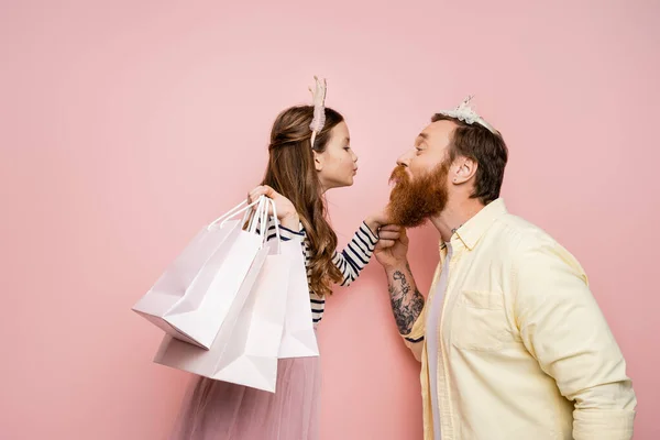 Vista lateral de la hija sosteniendo bolsas de compras y pucheros labios al padre con diadema de corona sobre fondo rosa - foto de stock