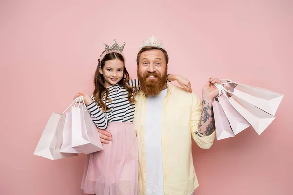 Alegre hombre e hija con diademas de corona sosteniendo bolsas de compras sobre fondo rosa - foto de stock