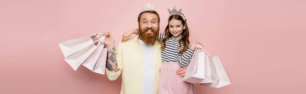 Happy father and girl with crown headbands holding shopping bags on pink background, banner — Stock Photo