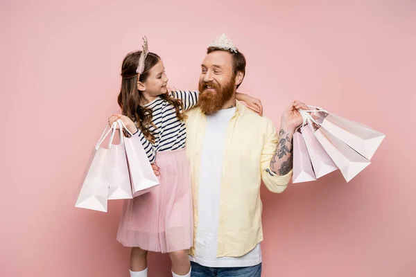 Chica alegre sosteniendo bolsas de compras y abrazando padre con diadema de corona sobre fondo rosa - foto de stock