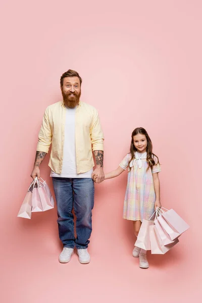 Full length of tattooed man holding hand of daughter and shopping bags on pink background — Stock Photo
