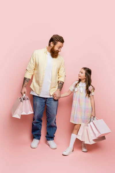 Веселый мужчина держит за руку дочь с пакетами для покупок на розовом фоне — стоковое фото