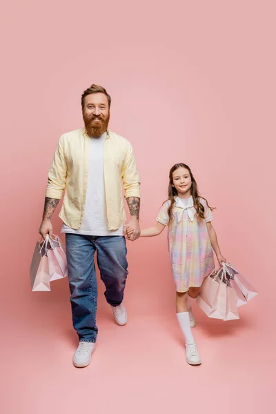 Улыбающиеся мужчина и дочь держат сумки с покупками во время прогулки на розовом фоне — стоковое фото