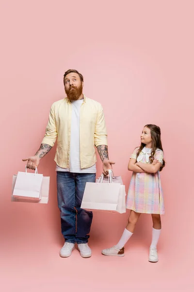 Preteen girl crossing arms near pensive father with shopping bags on pink background — Stock Photo