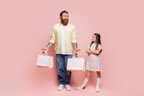 Full length of girl crossing arms near smiling dad with shopping bags on pink background — Stock Photo