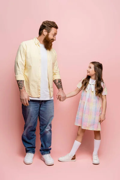 Вид сбоку улыбающегося татуированного мужчины, держащего за руку дочку-дочку на розовом фоне — стоковое фото