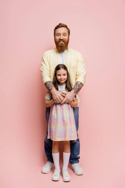 Повна довжина усміхненого татуйованого чоловіка обіймає дочку на рожевому фоні — стокове фото
