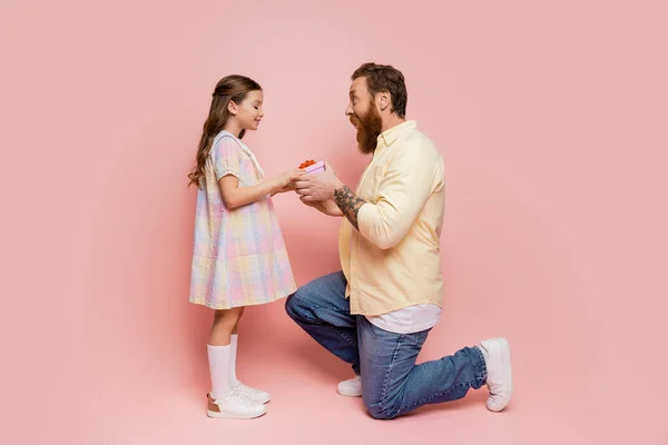Vista lateral del hombre tatuado dando regalo a la hija sonriente sobre fondo rosa - foto de stock