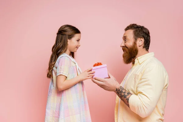 Вид збоку веселий бородатий чоловік дарує подарунок дочці ізольовано на рожевому — стокове фото