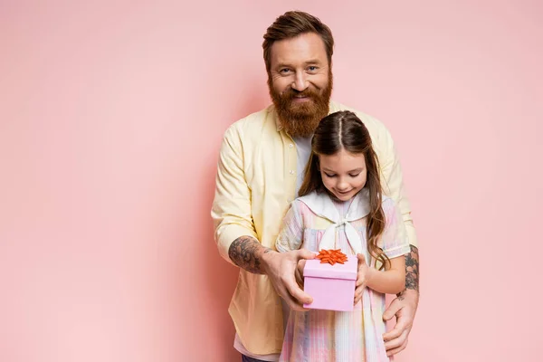 Улыбающийся бородатый мужчина дарит подарочную коробку дочери на розовом фоне — стоковое фото