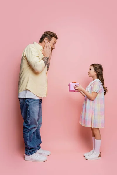 Vista lateral do homem chocado olhando para a filha pré-adolescente com presente no fundo rosa — Fotografia de Stock