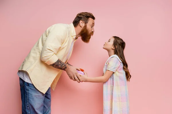 Side view of girl pouting lips while giving gift to bearded dad on pink background — Stock Photo