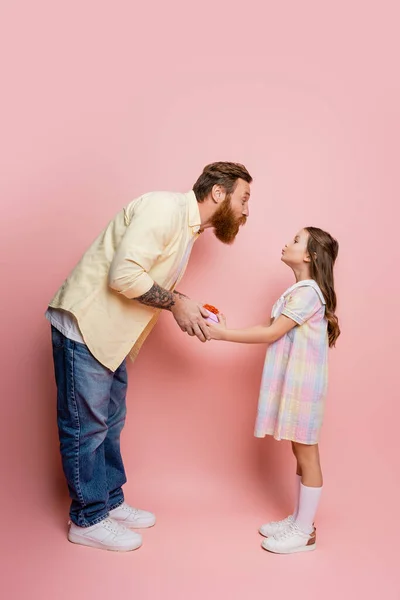 Vista lateral del hombre haciendo pucheros labios mientras le da caja de regalo a su hija sobre fondo rosa - foto de stock