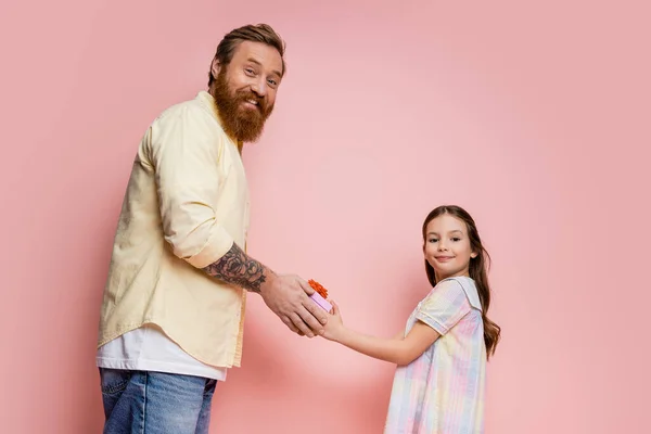 Fröhlicher bärtiger Vater und frühgeborenes Mädchen mit Geschenkschachtel und Blick in die Kamera auf rosa Hintergrund — Stockfoto