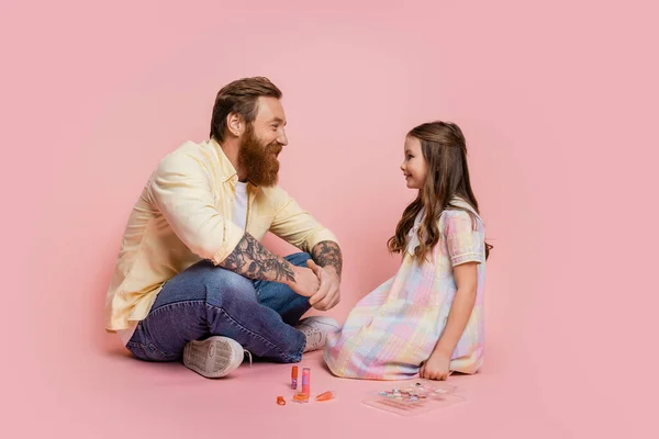 Vista lateral de sonriente padre tatuado sentado cerca de hija y cosméticos decorativos sobre fondo rosa - foto de stock