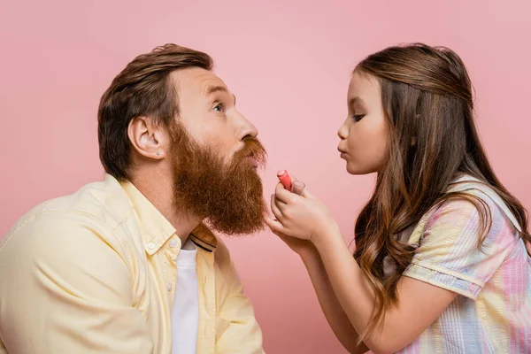 Chica sosteniendo lápiz labial y pucheros labios cerca de padre barbudo aislado en rosa - foto de stock