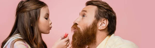 Ragazza preadolescente che tiene il rossetto vicino al padre barbuto isolato su rosa, banner — Foto stock